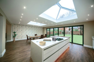 Interior of a bespoke orangery in Somerset with wide patio doors leading to a garden. There is a dining table, and kitchen island in the foreground and two large skylights and ceiling spotlights.